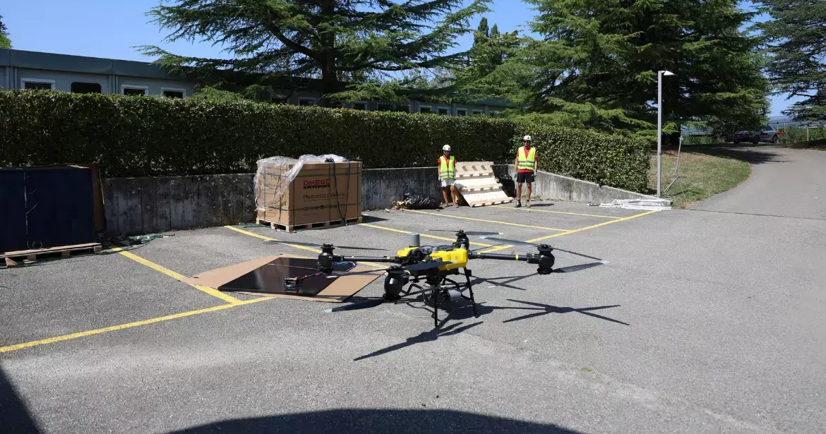 Drone de transport au sol devant un panneau solaire prêt à être levé, avec deux techniciens en gilet de sécurité en arrière-plan.