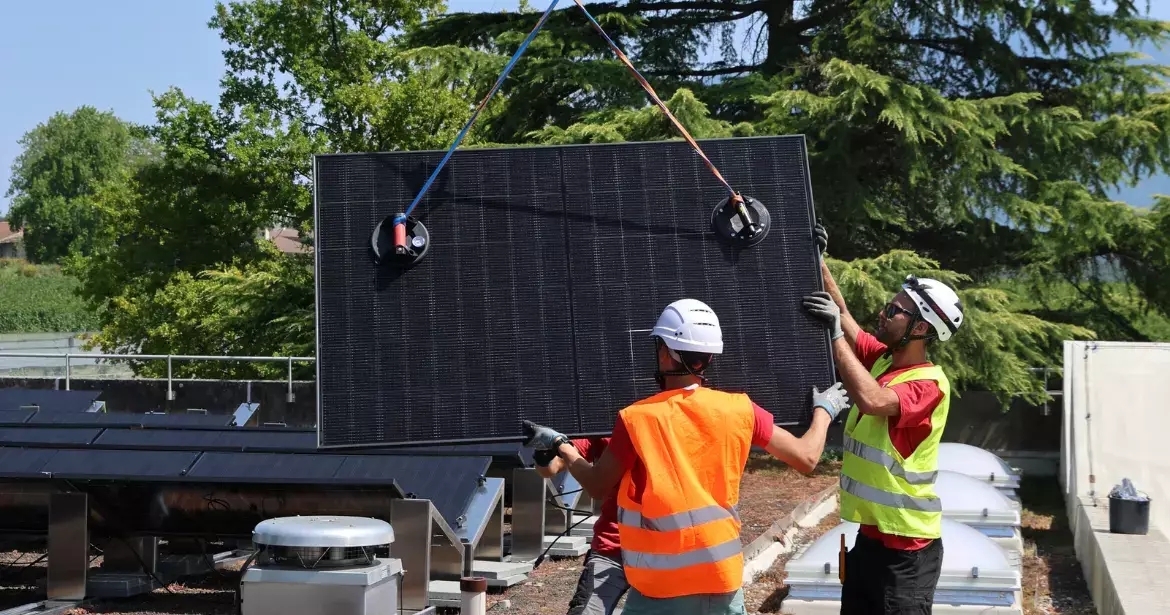 Deux techniciens installent un panneau photovoltaïque suspendu par des sangles, sur l’un des toitures de l’établissement.