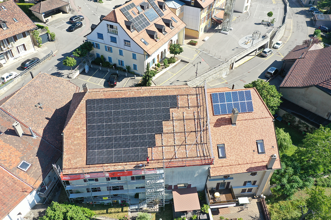 Vue aérienne de panneaux solaires sur plusieurs toits en terre cuite d'un village, avec échafaudage visible sur un bâtiment principal.