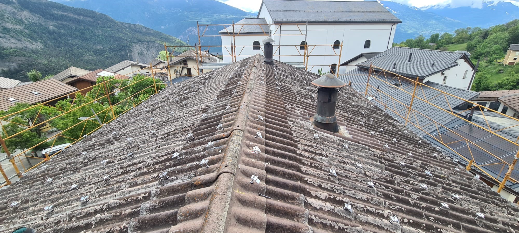 Toiture en tuiles avec supports de fixation pour panneaux solaires photovoltaïques, entourée d’un échafaudage de sécurité, dans le village de Ayen en Valais avec vue sur les Alpes valaisannes. Préparation de l’installation solaire sur toit incliné, en environnement résidentiel alpin.