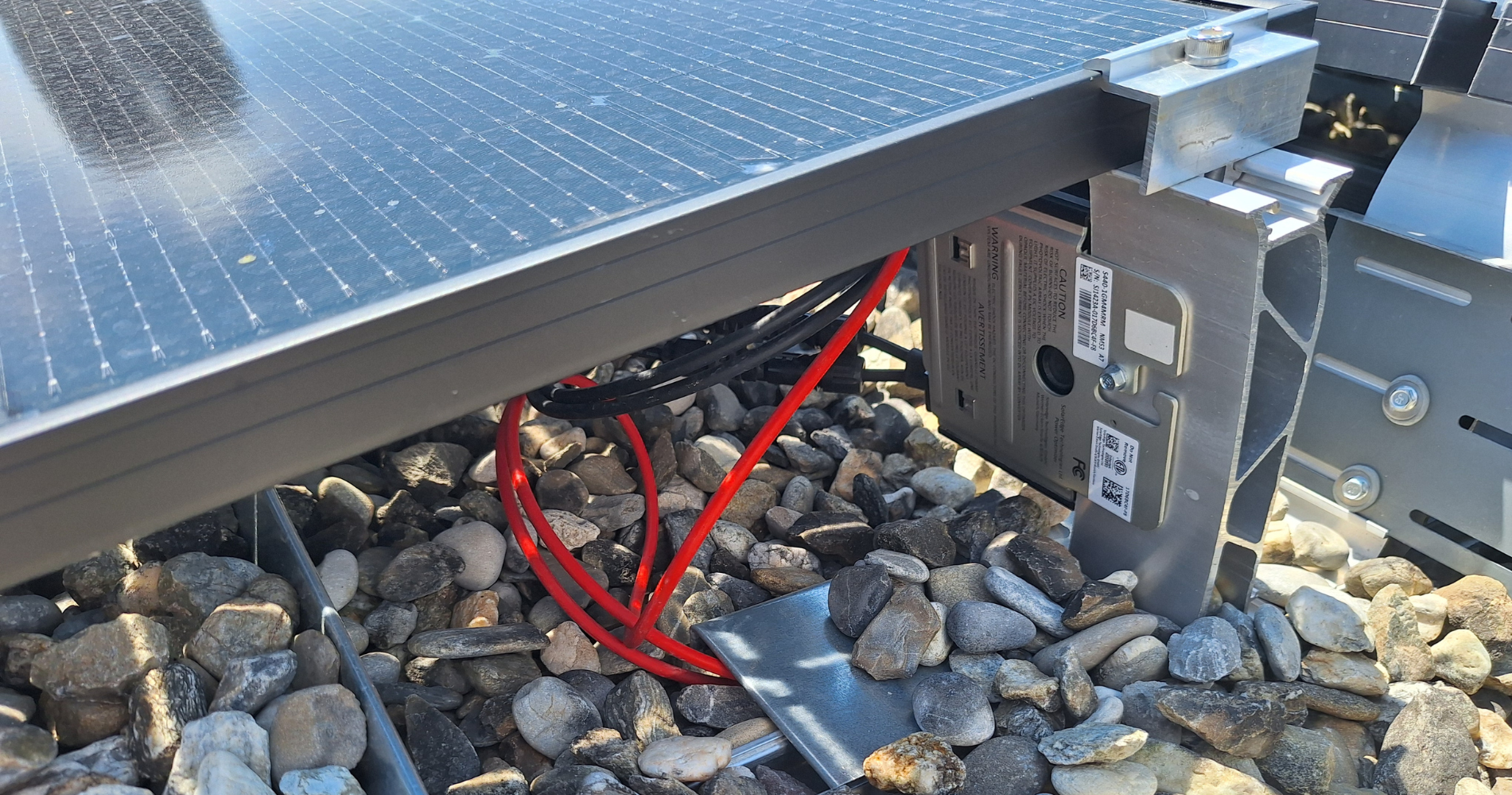 Vue rapprochée sous un panneau photovoltaïque montrant les câbles électriques et les éléments de fixation posés sur une toiture plate en gravier.